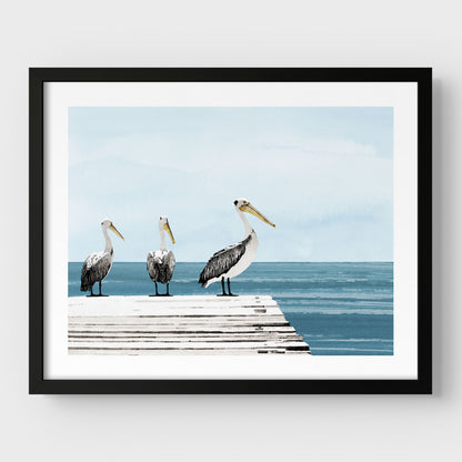 Black framed illustration of three pelicans standing on a weathered dock above calm blue ocean water, with a soft, pale blue sky in the background.
