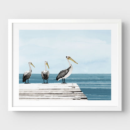White framed illustration of three pelicans standing on a weathered dock above calm blue ocean water, with a soft, pale blue sky in the background.
