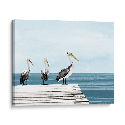 Canvas print of three pelicans standing on a sunlit dock overlooking calm blue ocean water, with a soft gradient sky in the background.