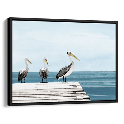 Black framed canvas print featuring three pelicans standing on a weathered white dock above calm blue ocean water, under a soft, cloudy sky.