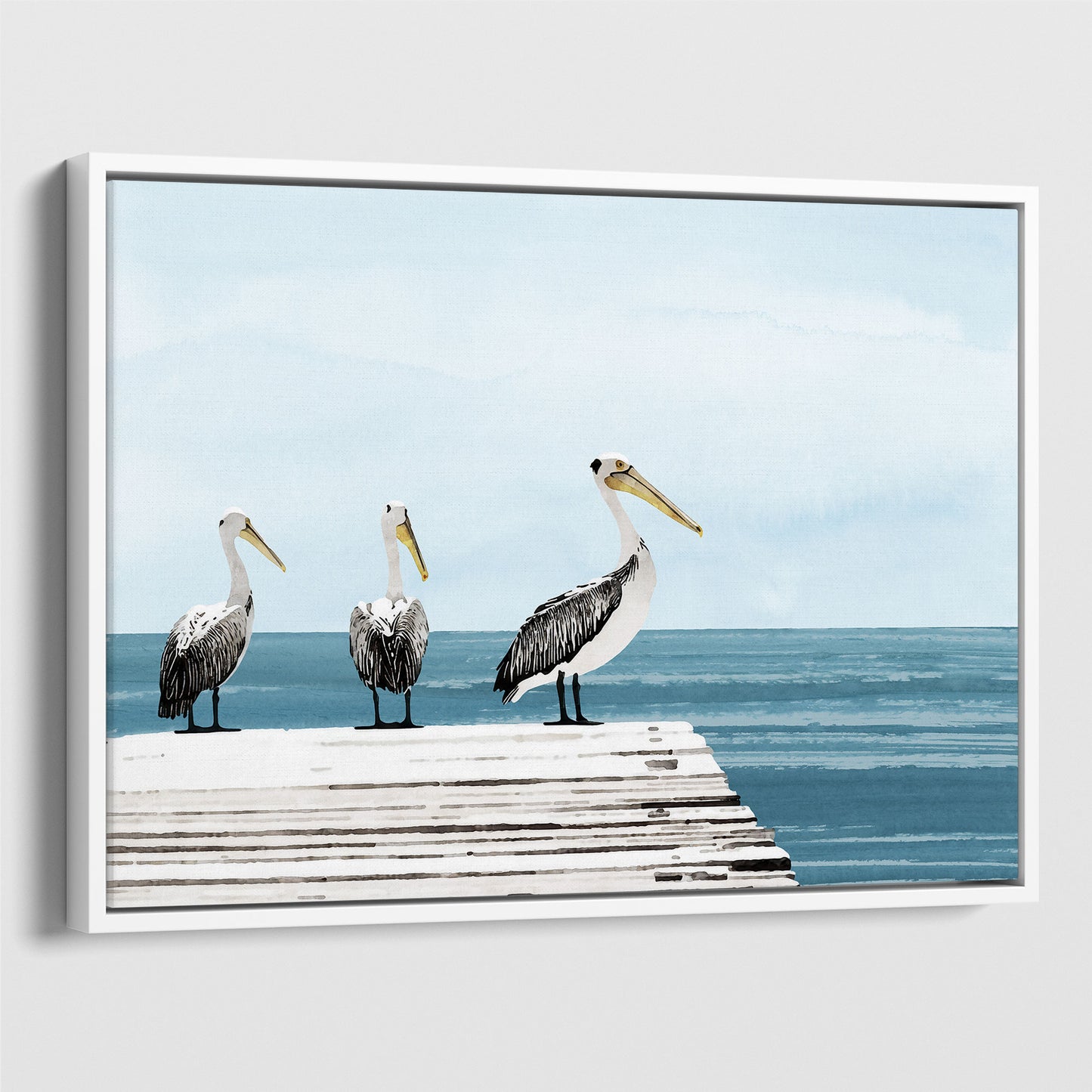 White framed canvas print featuring three pelicans standing on a weathered white dock above calm blue ocean water, under a soft, cloudy sky.