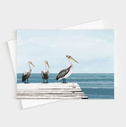 Illustrated card showing three pelicans standing on a weathered white dock overlooking a calm, blue ocean with a soft, pale sky in the background.