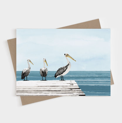 Illustrated card showing three pelicans standing on a weathered white dock overlooking a calm, blue ocean with a soft, pale sky in the background.