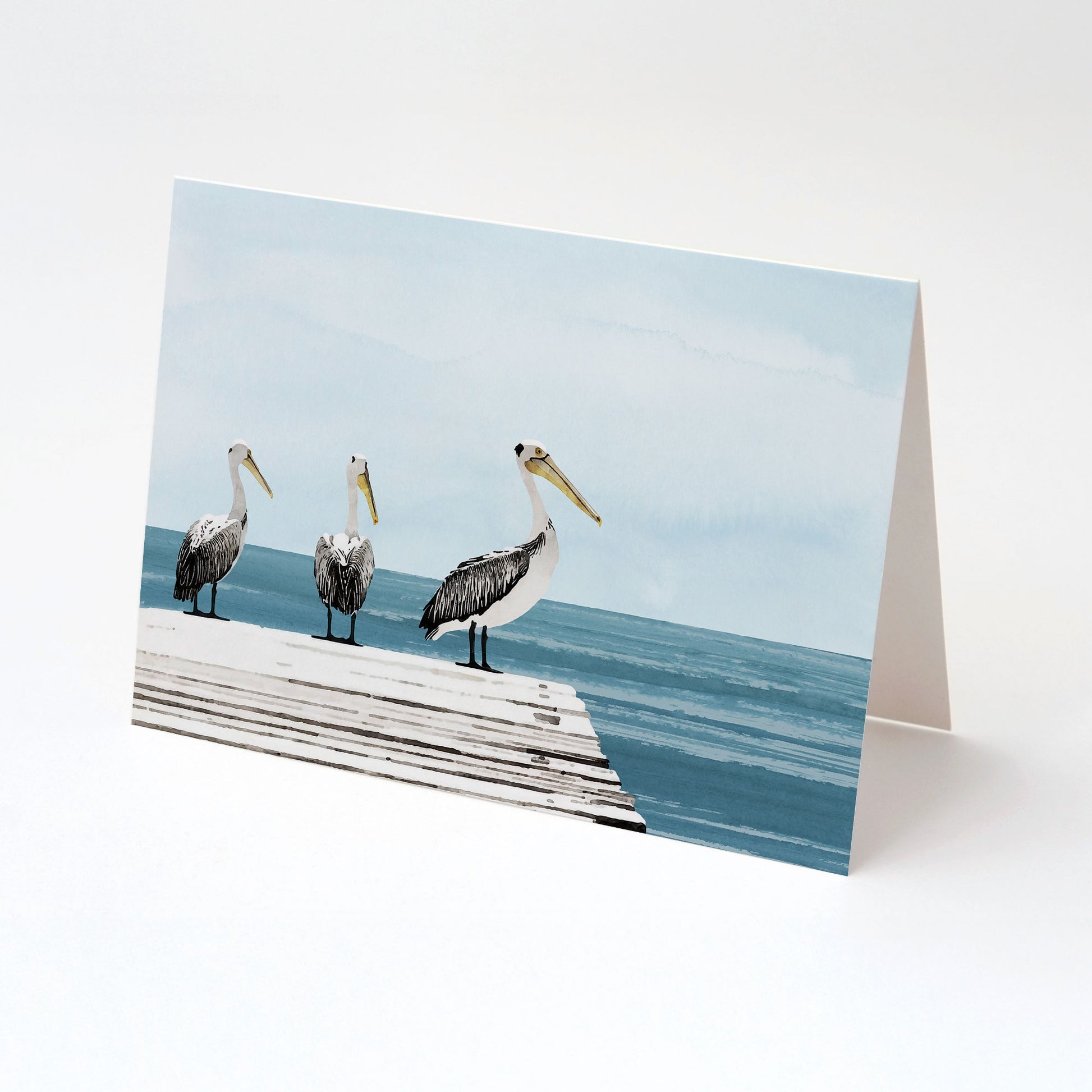 Folded greeting card displaying a watercolor-style illustration of three pelicans standing on a wooden dock beside a calm blue ocean, set against a pale blue sky with soft gradients.