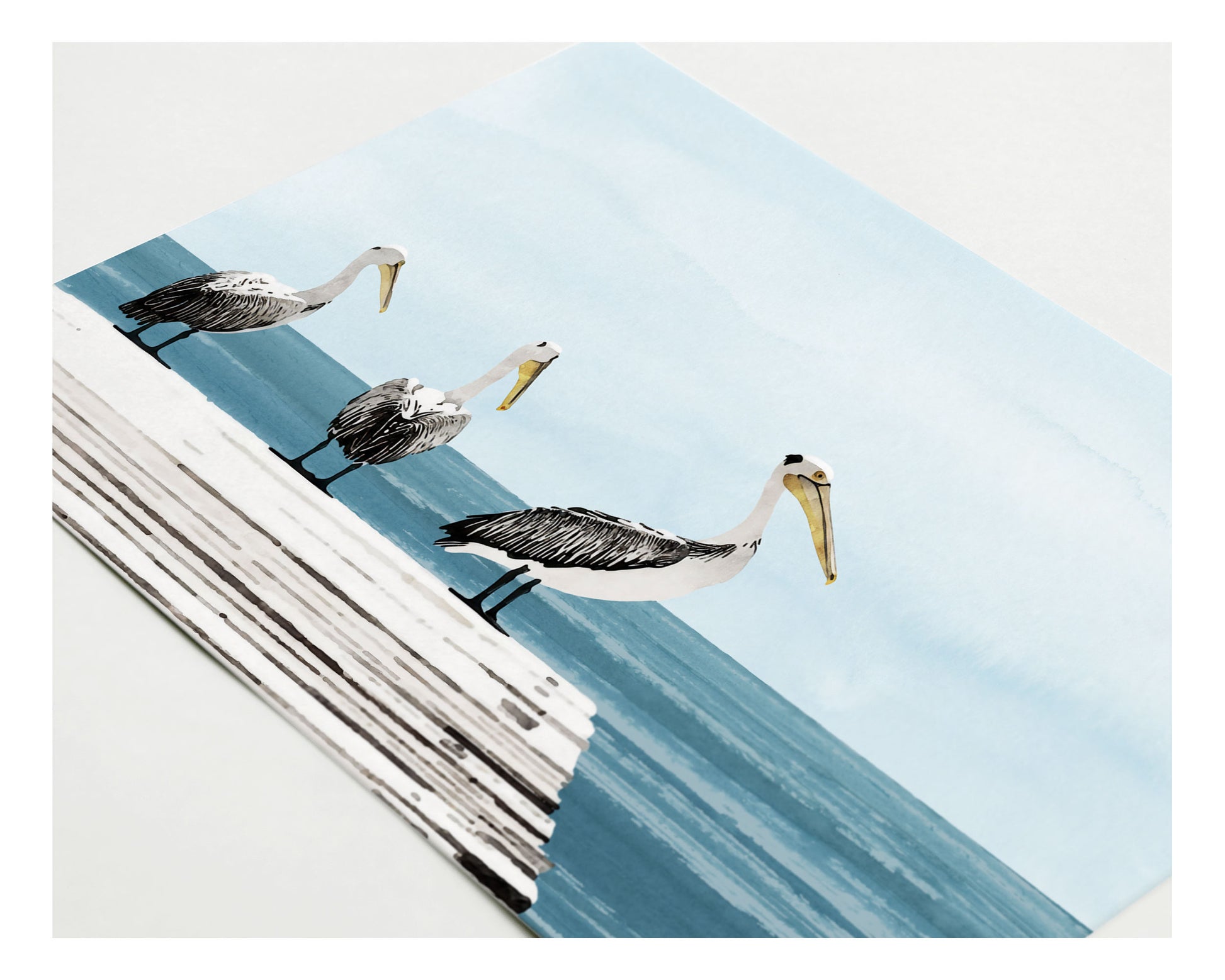 Close-up of a greeting card featuring a watercolor-style illustration of three pelicans standing on a wooden dock above a calm blue sea, under a soft, pale sky.