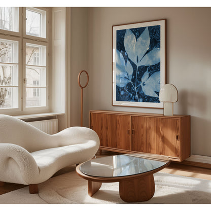 Framed cyanotype-style print of peony leaves in soft blue tones, hanging above a wood sideboard in a modern, light-filled living room with curved white seating and minimalist decor.