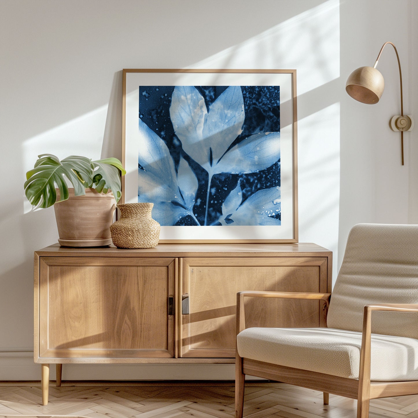 Framed botanical cyanotype of peony leaves in deep blue and white tones, leaning on a wood sideboard in a sunlit room with minimalist decor, a woven jug, a potted monstera, and a neutral lounge chair.