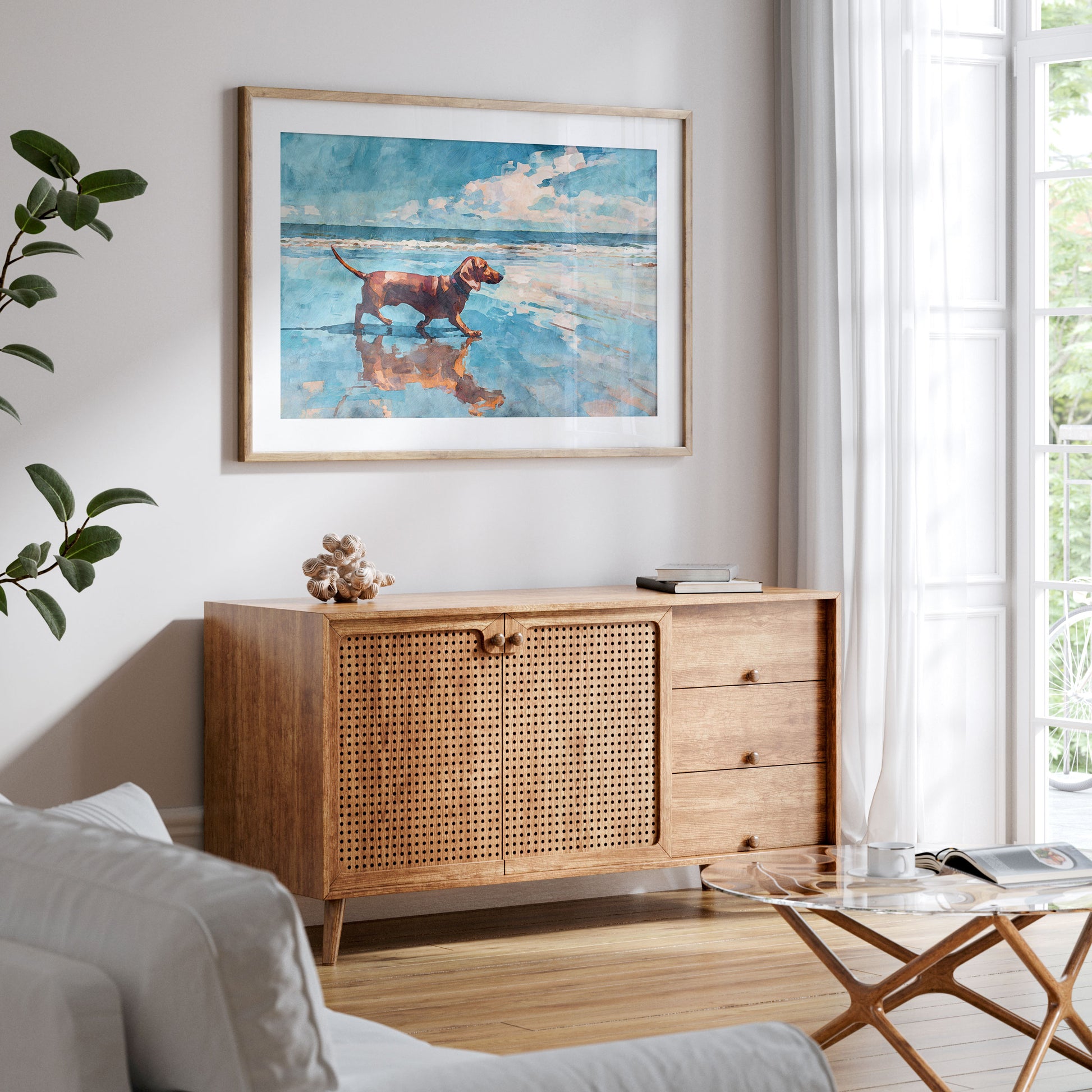 Framed watercolor-style beach scene of a red dachshund walking along the shoreline, reflected in wet sand under a bright blue sky. Displayed above a mid-century wood sideboard with cane doors in a light, airy living room with neutral tones and soft natural light.