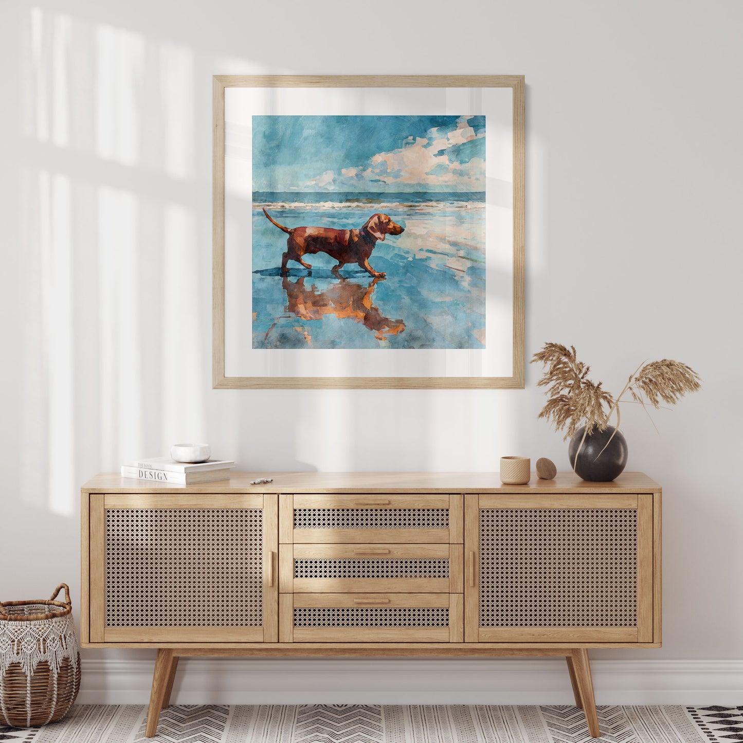 Framed watercolor-style painting of a red dachshund walking along a beach shoreline, reflected in wet sand beneath a blue sky with soft clouds. Displayed above a light wood cane-front sideboard with neutral decor, warm sunlight casting soft shadows across a calm, minimalist interior.