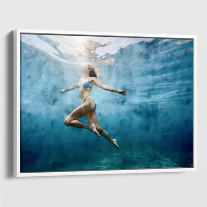 view of a woman in a blue swimsuit swimming upward, surrounded by blue-green water with light filtering from above. Artwork printed on gallery wrap canvas in white floater frame.