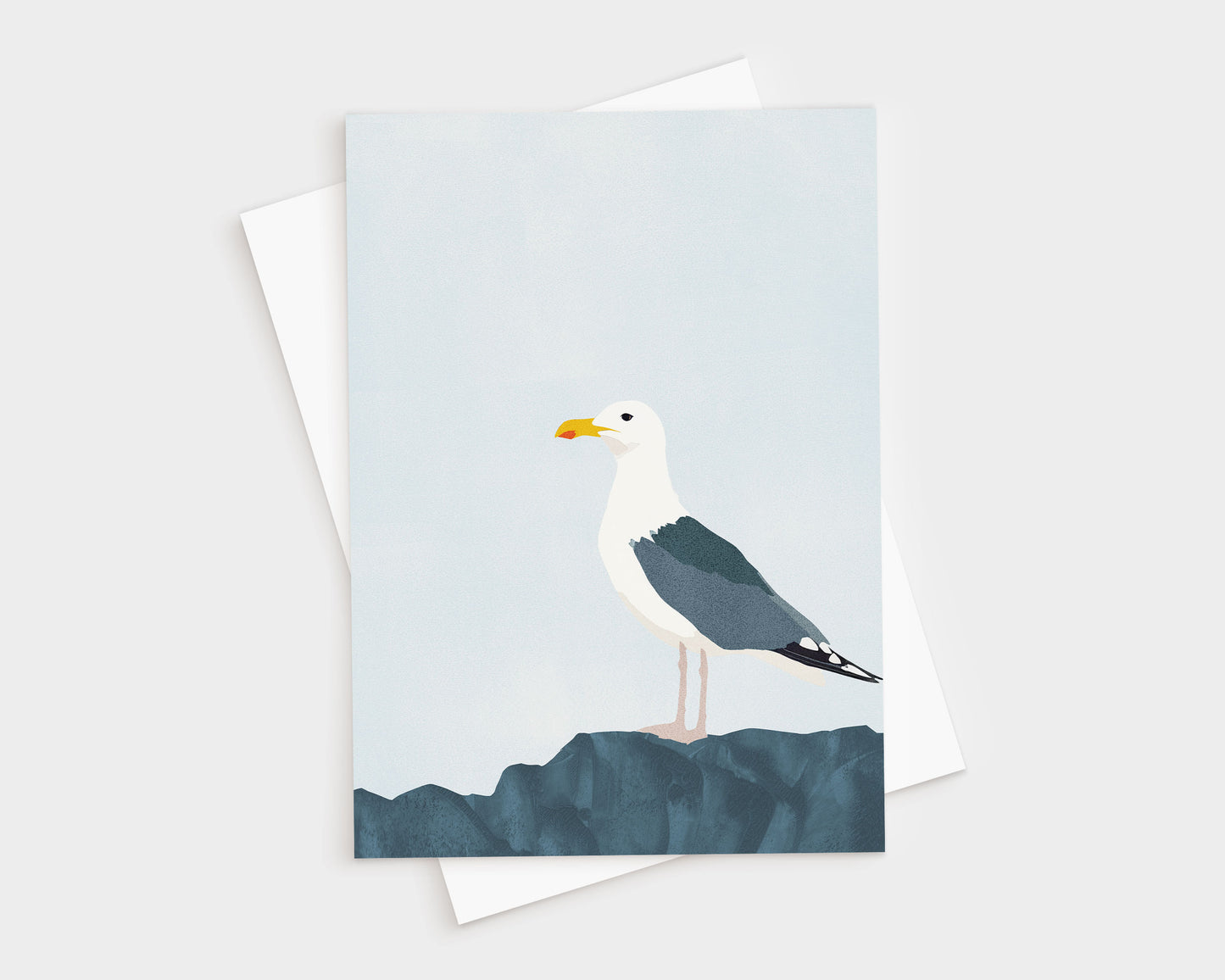 Minimalist greeting card featuring a standing seagull with dark wings and white head perched on textured blue rocks, set against a pale blue sky, with a white envelope underneath.