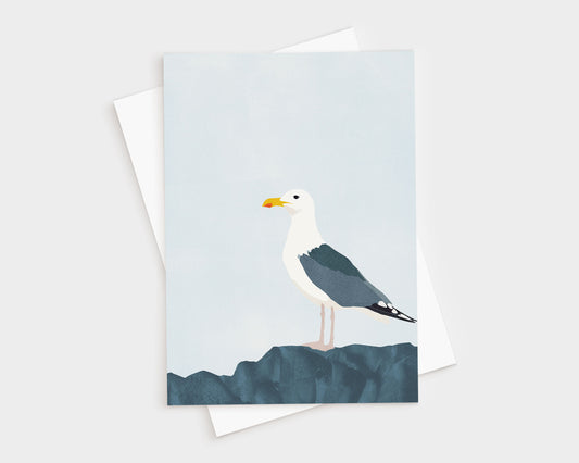 Minimalist greeting card featuring a standing seagull with dark wings and white head perched on textured blue rocks, set against a pale blue sky, with a white envelope underneath.