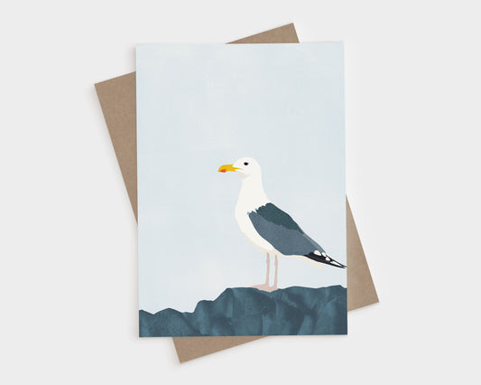 Minimalist greeting card featuring a standing seagull with dark wings and white head perched on textured blue rocks, set against a pale blue sky, with a kraft brown envelope underneath.
