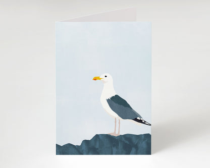 Minimalist greeting card standing upright, featuring a seagull with dark wings and white head perched on textured blue rocks beneath a pale sky.