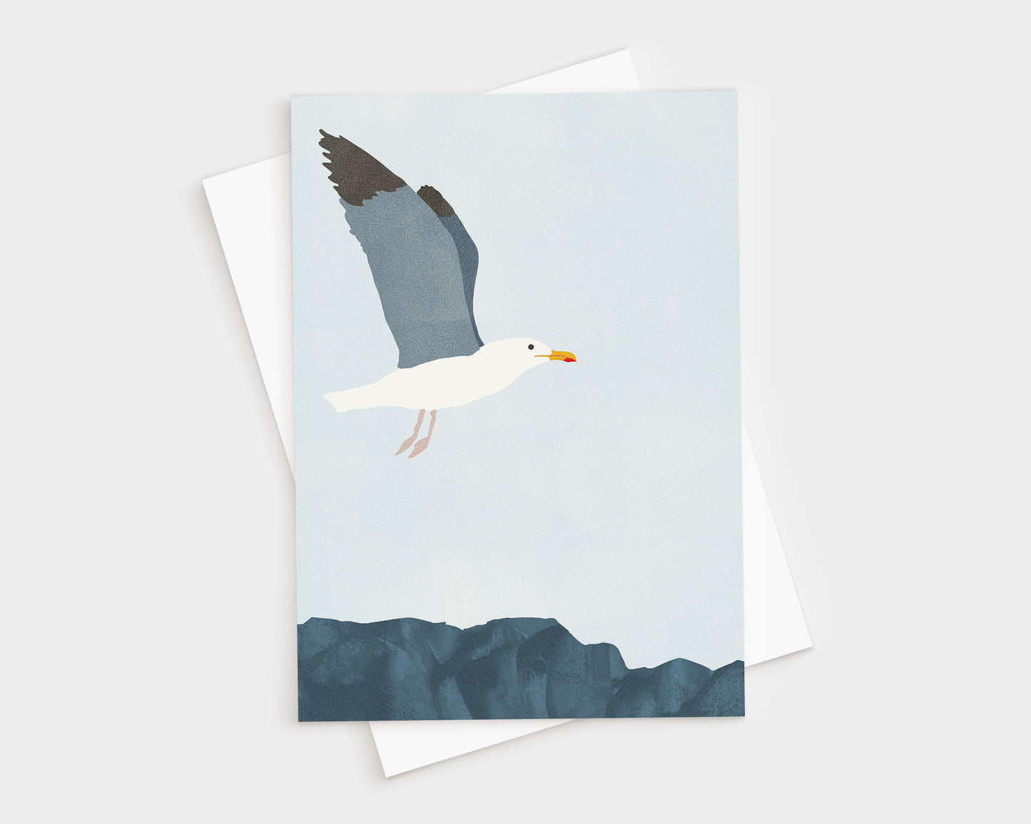 Minimalist illustration of a seagull in flight above dark blue coastal rocks, set against a pale blue sky, featured on a greeting card with a white envelope behind it.