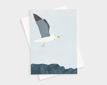 Minimalist illustration of a seagull in flight above dark blue coastal rocks, set against a pale blue sky, featured on a greeting card with a white envelope behind it.