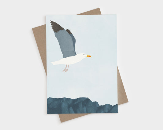 Minimalist illustration of a seagull in flight above dark blue coastal rocks, set against a pale blue sky, featured on a greeting card with a kraft brown envelope behind it.