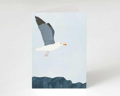 Minimalist greeting card standing upright, featuring a white seagull with dark wings flying over deep blue rocks against a pale blue sky.