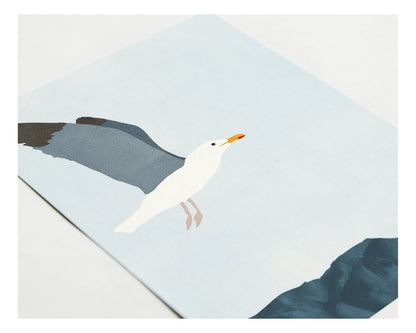 Close-up of a greeting card with a minimalist illustration of a seagull flying over dark blue rocks against a pale sky, highlighting the texture and detail of the printed design.