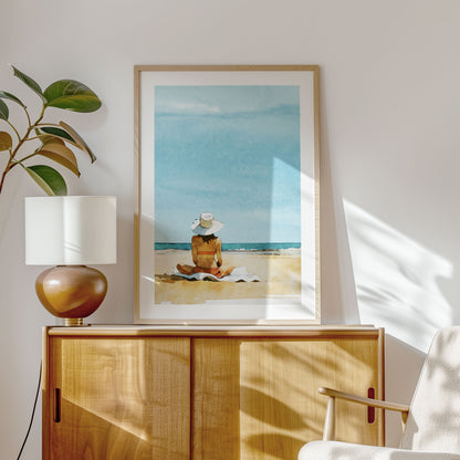 Framed artwork of a person sitting on a beach, displayed on a wooden cabinet with a table lamp and a potted plant, in a bright room with soft natural light.