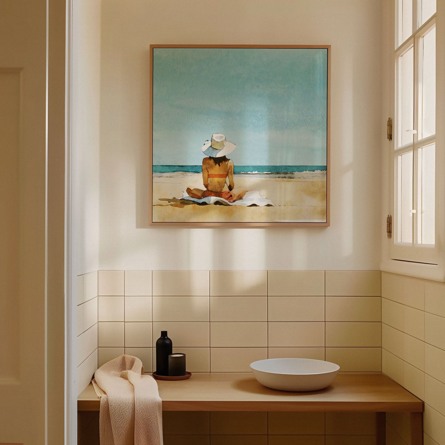 Framed beach artwork hanging above a wooden countertop with a ceramic sink, folded towel, and decor items, in a softly lit bathroom with tiled walls and a window.