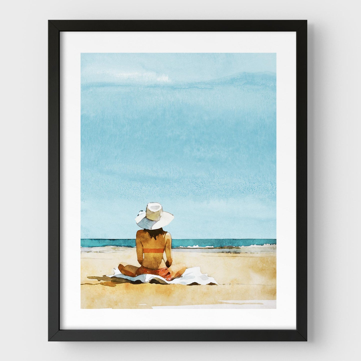 Black framed artwork featuring a person sitting on a towel at a beach, wearing a wide-brimmed white hat and a swimsuit, with a clear blue sky and ocean in the background.