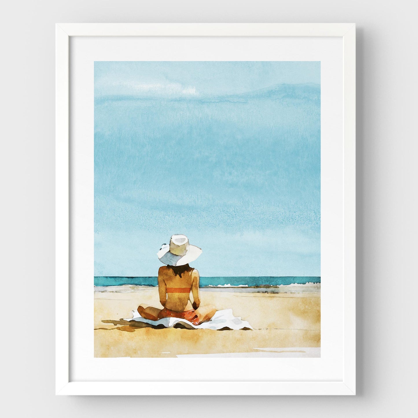 White framed artwork featuring a person sitting on a towel at a beach, wearing a wide-brimmed white hat and a swimsuit, with a clear blue sky and ocean in the background.