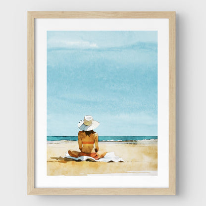 Natural wood framed artwork featuring a person sitting on a towel at a beach, wearing a wide-brimmed white hat and a swimsuit, with a clear blue sky and ocean in the background.