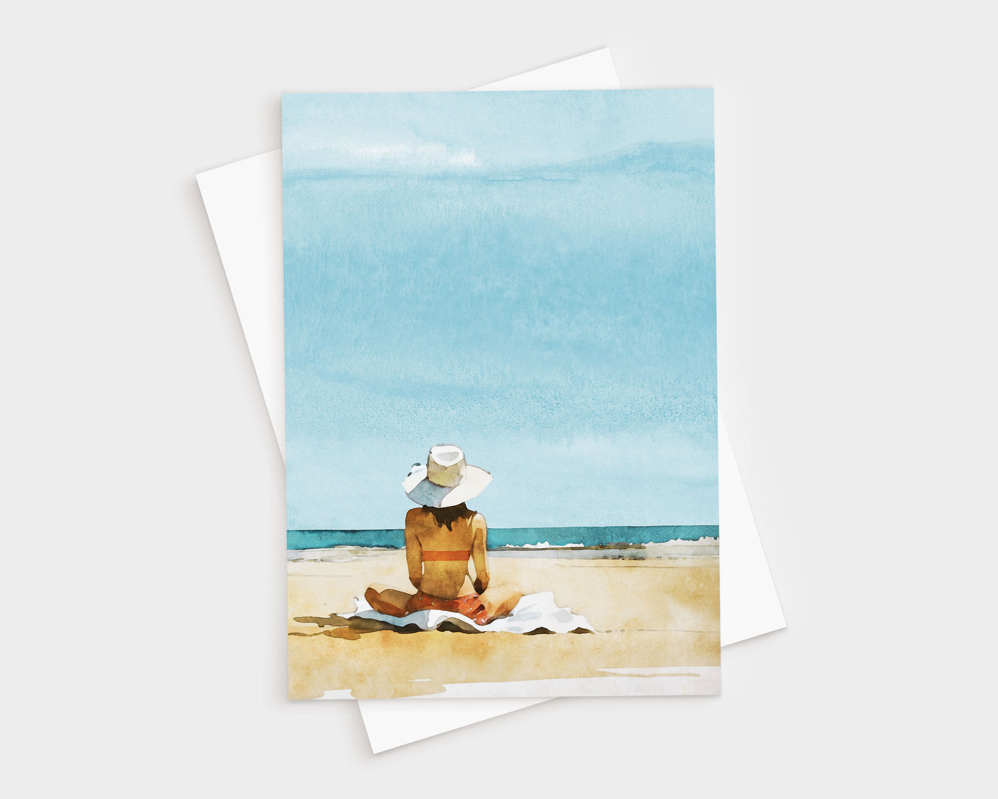 Greeting card featuring a watercolor-style illustration of a woman in a sunhat and swimsuit sitting on a towel at the beach, facing the ocean under a bright blue sky.