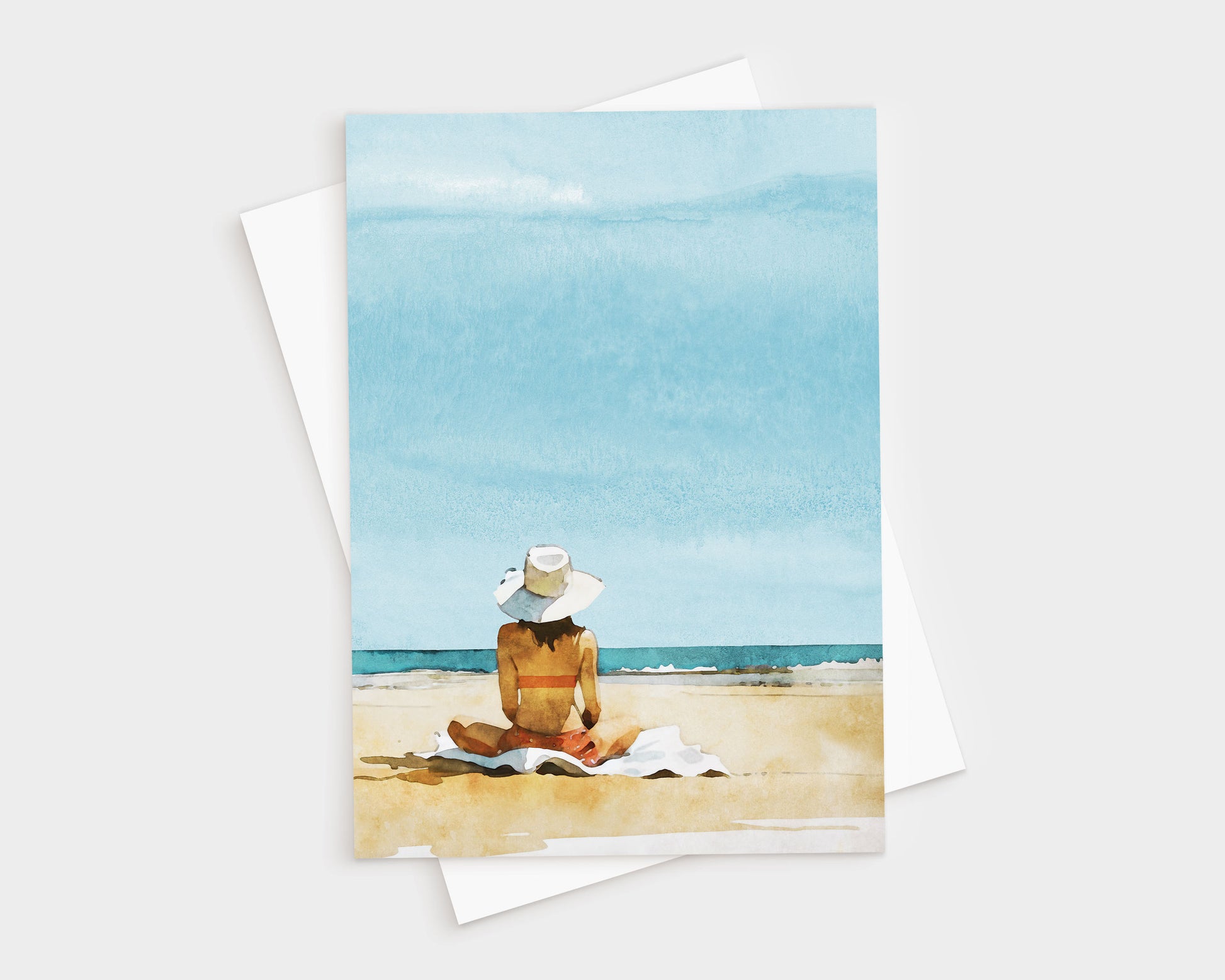 Greeting card featuring a watercolor-style illustration of a woman in a sunhat and swimsuit sitting on a towel at the beach, facing the ocean under a bright blue sky.