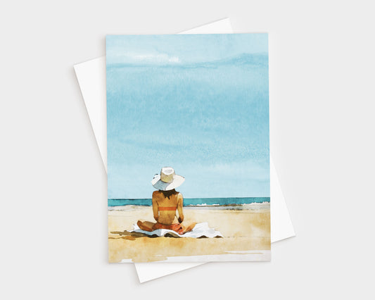Greeting card featuring a watercolor-style illustration of a woman in a sunhat and swimsuit sitting on a towel at the beach, facing the ocean under a bright blue sky.