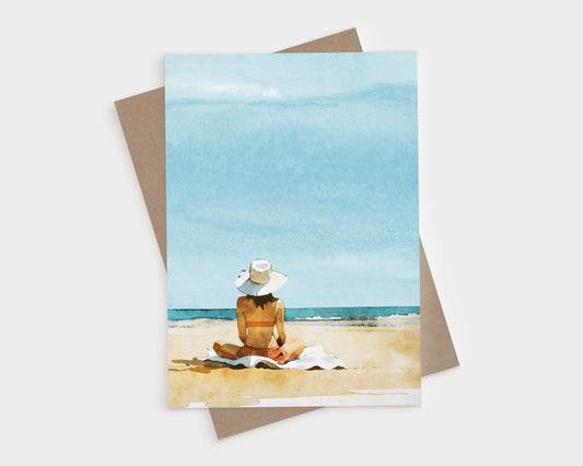 Greeting card with kraft envelope featuring a watercolor-style illustration of a woman in a sunhat and swimsuit sitting on a towel at the beach, facing the ocean under a bright blue sky.