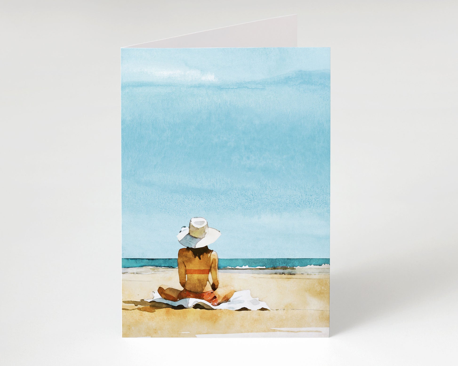 Minimalist beach-themed greeting card shown upright, featuring a watercolor illustration of a woman in a sunhat and bikini sitting on a towel, looking out at the ocean under a soft blue sky.