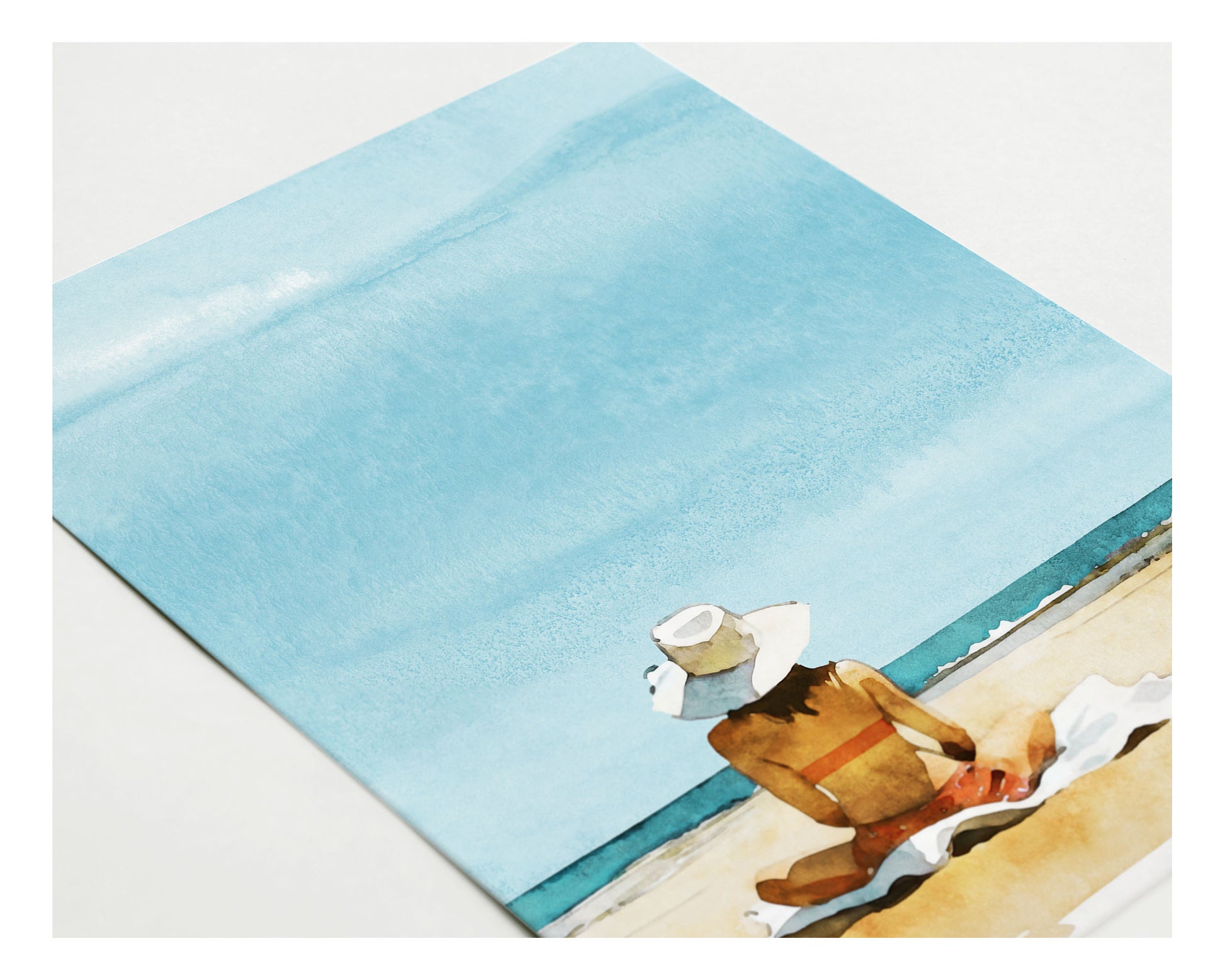 Close-up angled view of a greeting card with a watercolor-style illustration of a woman in a sunhat sitting on a towel at the beach, gazing at the ocean under a soft blue sky.