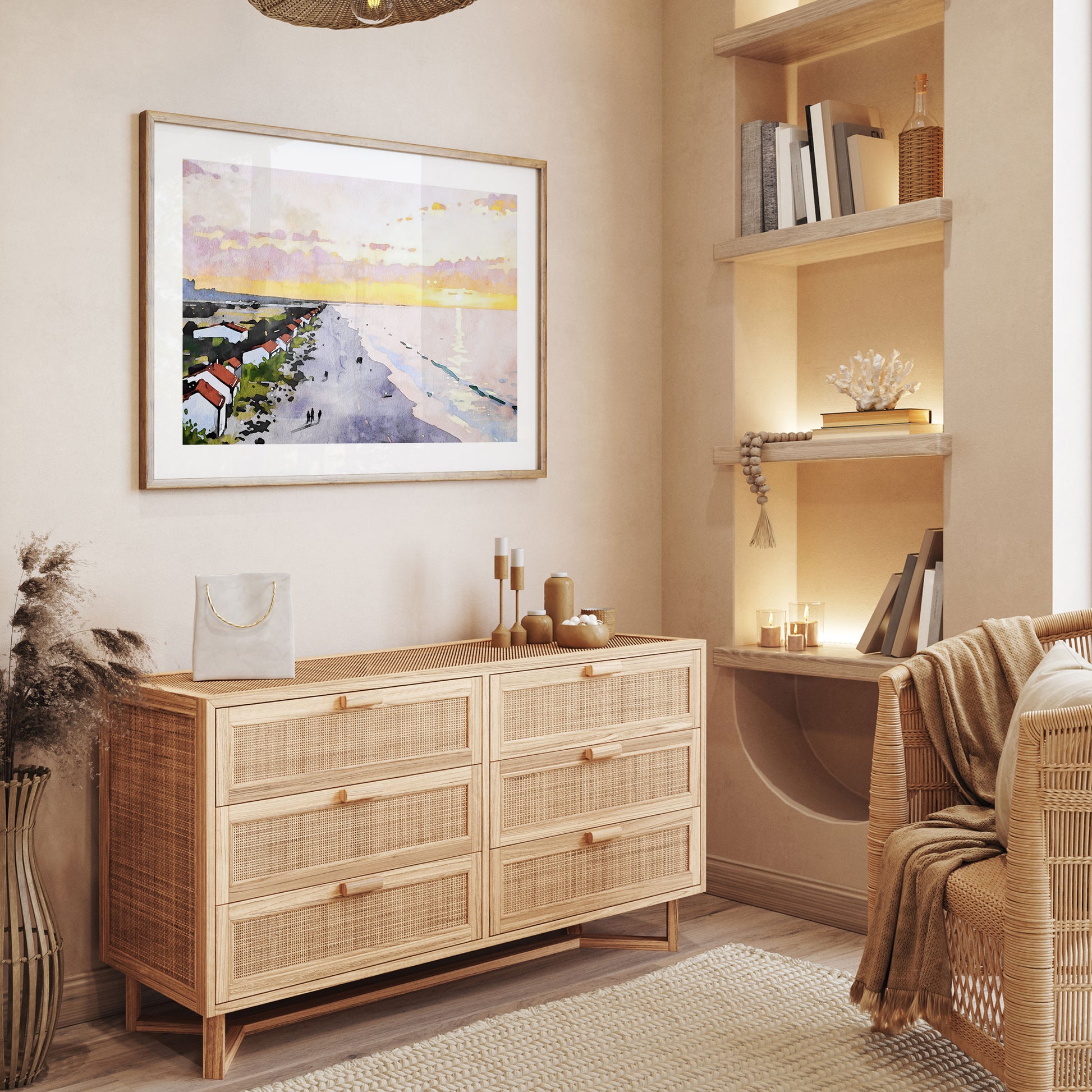 Framed artwork displayed in a cozy, neutral-toned room, featuring a watercolor-style depiction of a sunrise beach scene with soft pastel skies and a reflective ocean. The artwork is mounted above a natural wooden dresser.