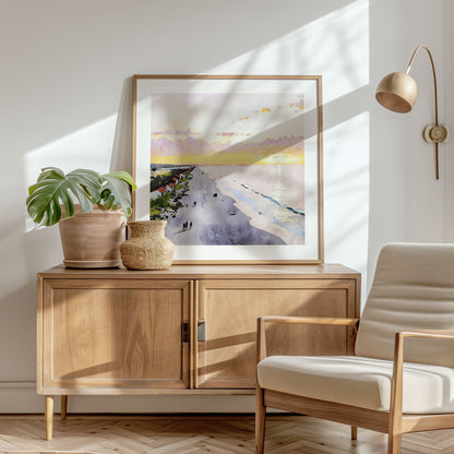 Framed artwork in a natural wood frame, leaning on a wooden console in a bright, sunlit room, depicting a watercolor-style sunrise beach scene. The console is styled with a potted plant and a woven basket.