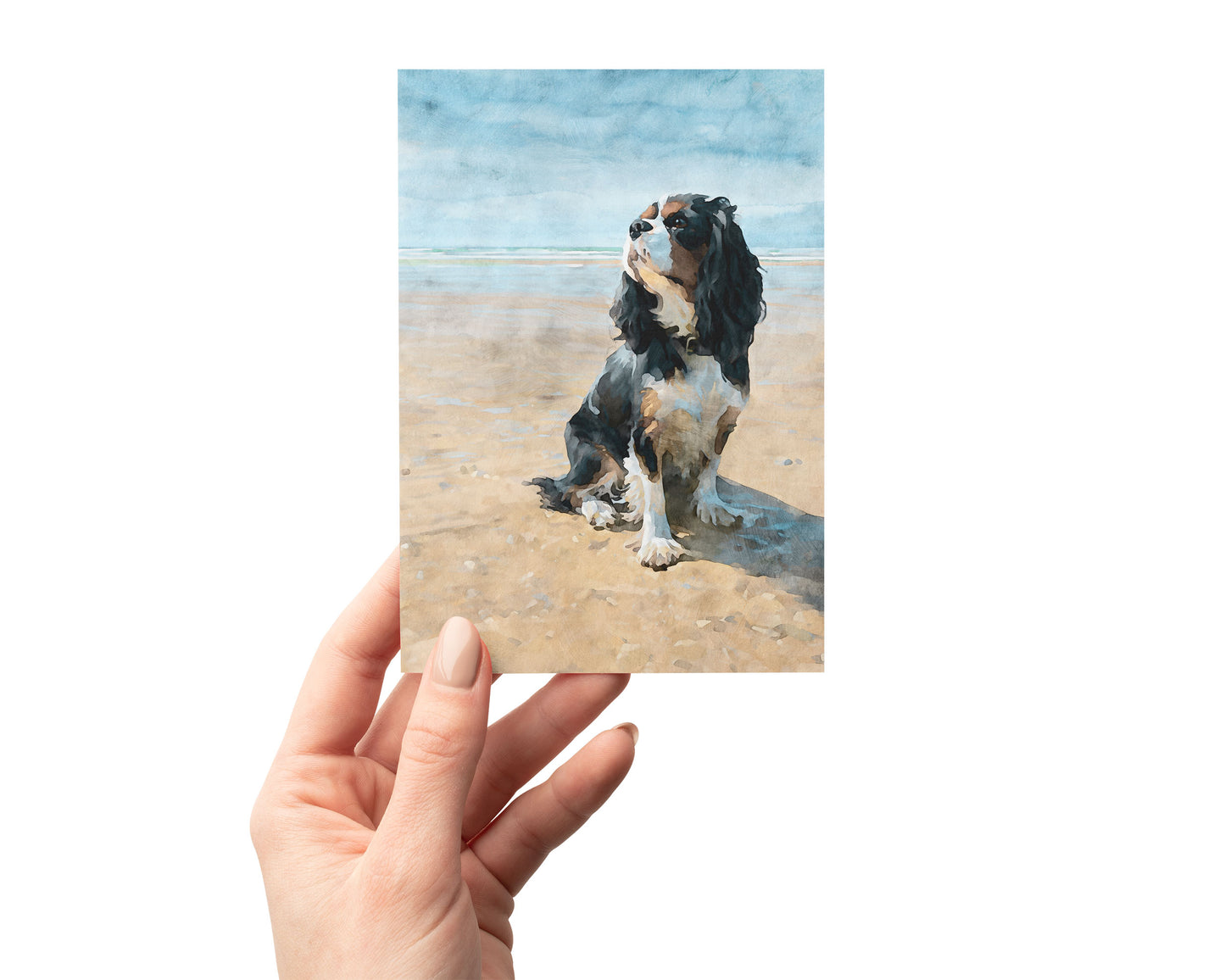 Hand holding a card with a tricolor Cavalier King Charles spaniel painting on a beach