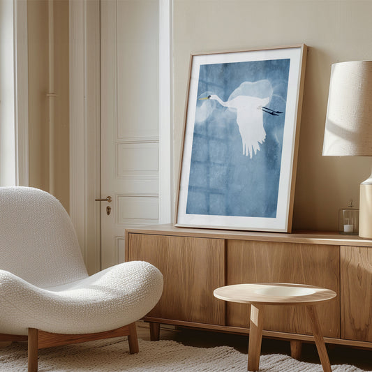 Framed minimalist print of a white heron flying with wings down, displayed in a cozy, sunlit room with neutral tones, wood furniture, and a soft textured chair.