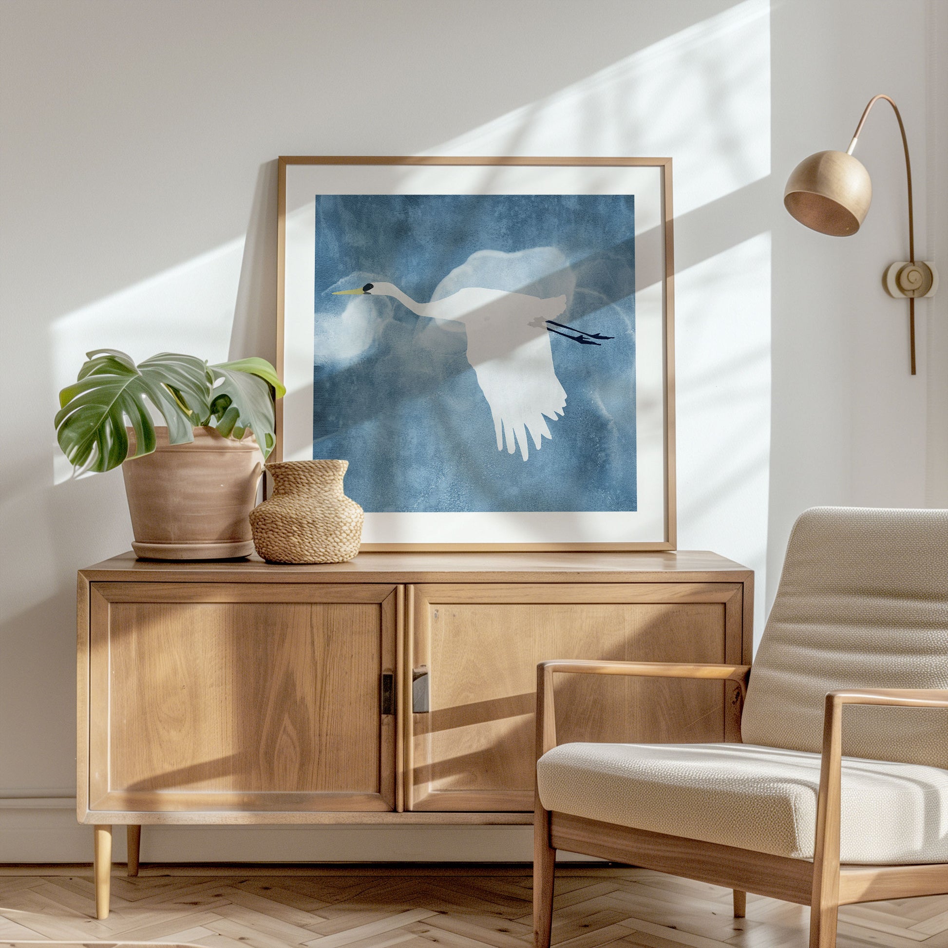 Framed square print of a white heron in flight with wings lowered, displayed in a bright, minimalist room with wooden furniture, a woven vase, and a potted monstera, bathed in natural light.