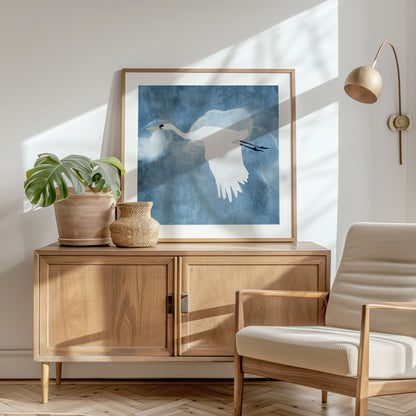Framed square print of a white heron in flight with wings lowered, displayed in a bright, minimalist room with wooden furniture, a woven vase, and a potted monstera, bathed in natural light.