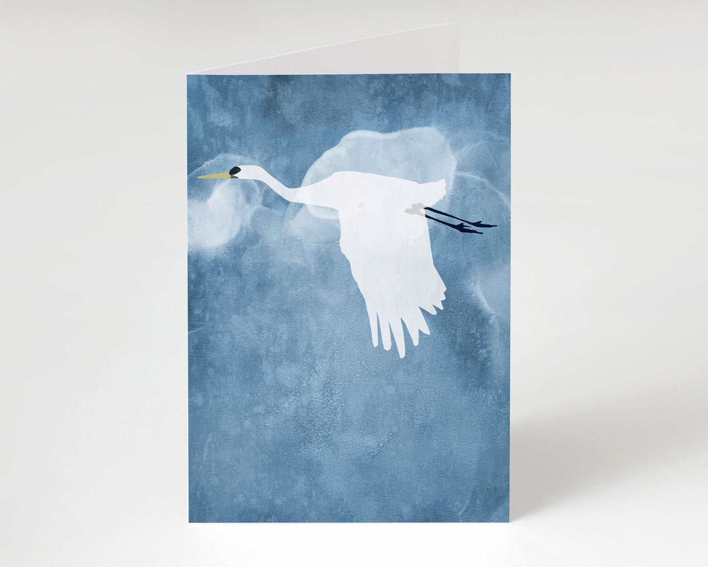 Greeting card featuring a white heron in flight against a textured, cloud-like blue background, shown standing upright with a slight shadow on a plain white surface.