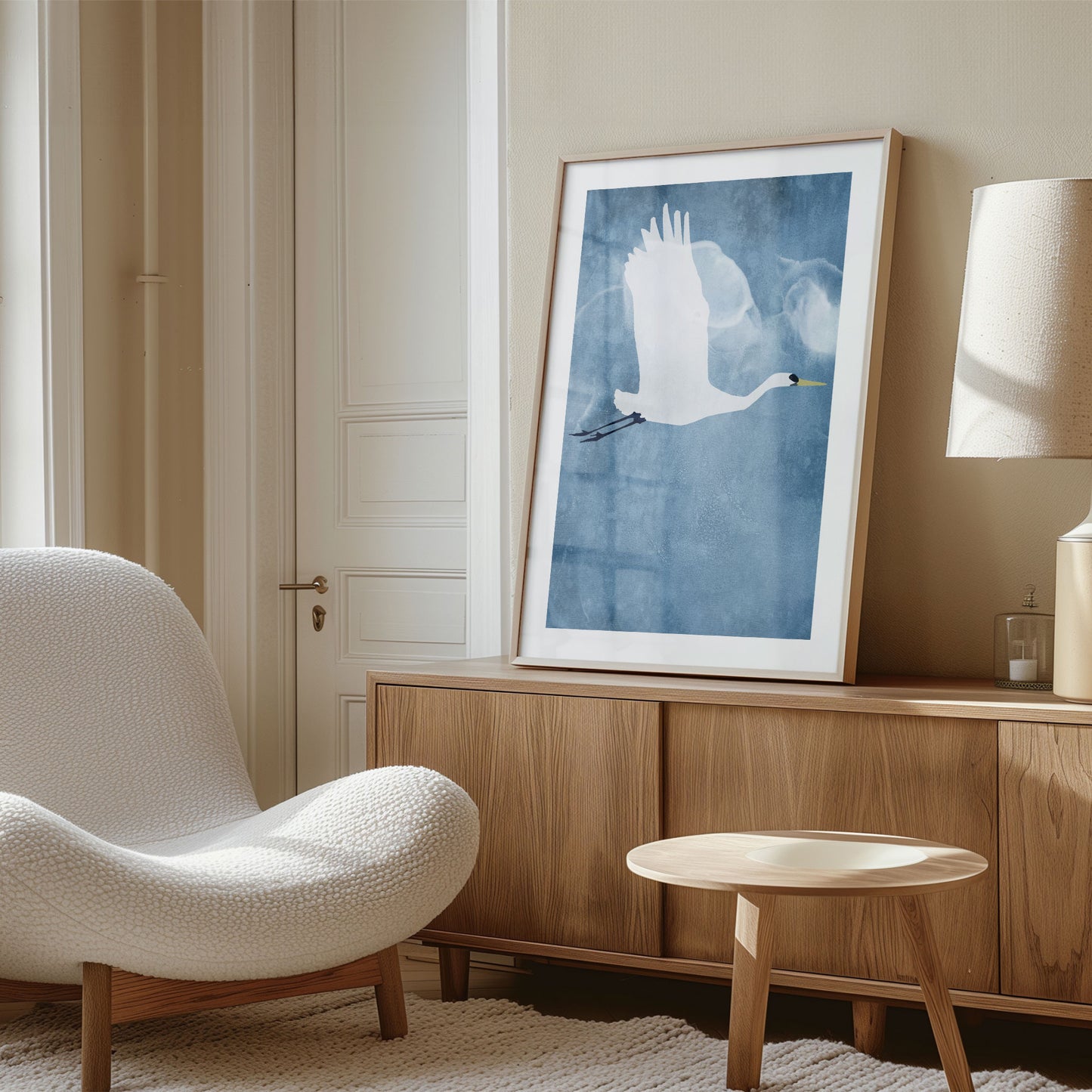 Framed print of a white heron in flight with wings lifted, displayed on a wood sideboard in a sunlit, modern living space with neutral tones and minimalist decor.