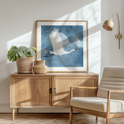 Framed square print of a white heron flying against a blue textured background, displayed on a mid-century wood sideboard with a potted monstera, woven vase, and soft natural light in a cozy, modern interior.