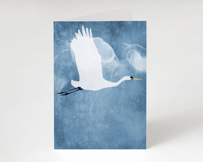 Greeting card showing a minimalist white heron with wings raised in flight, set against a moody, cloud-textured blue background.