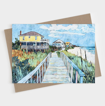 Folded greeting card featuring a vibrant watercolor-style illustration of a coastal boardwalk leading past colorful beach houses toward the ocean. A bright yellow house stands out in the foreground, framed by sea oats, greenery, and soft blue skies, capturing the relaxed charm of a seaside neighborhood.
