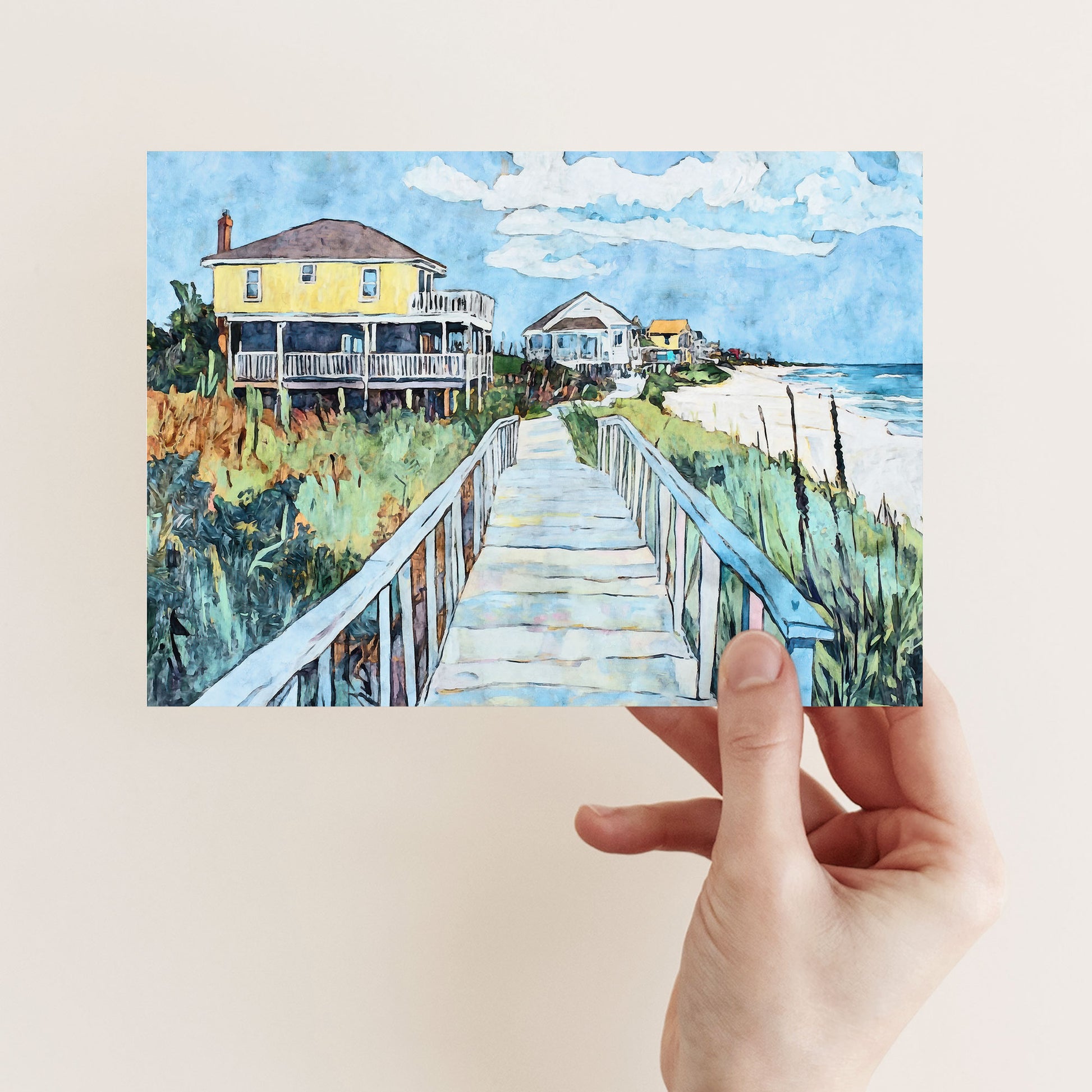 A hand holds a folded greeting card featuring a vivid watercolor-style beach scene. A wooden boardwalk leads through sea grass toward pastel-colored coastal houses, with a bright yellow house prominently on the left. The ocean waves and a clear blue sky with fluffy clouds create a peaceful, summery atmosphere.