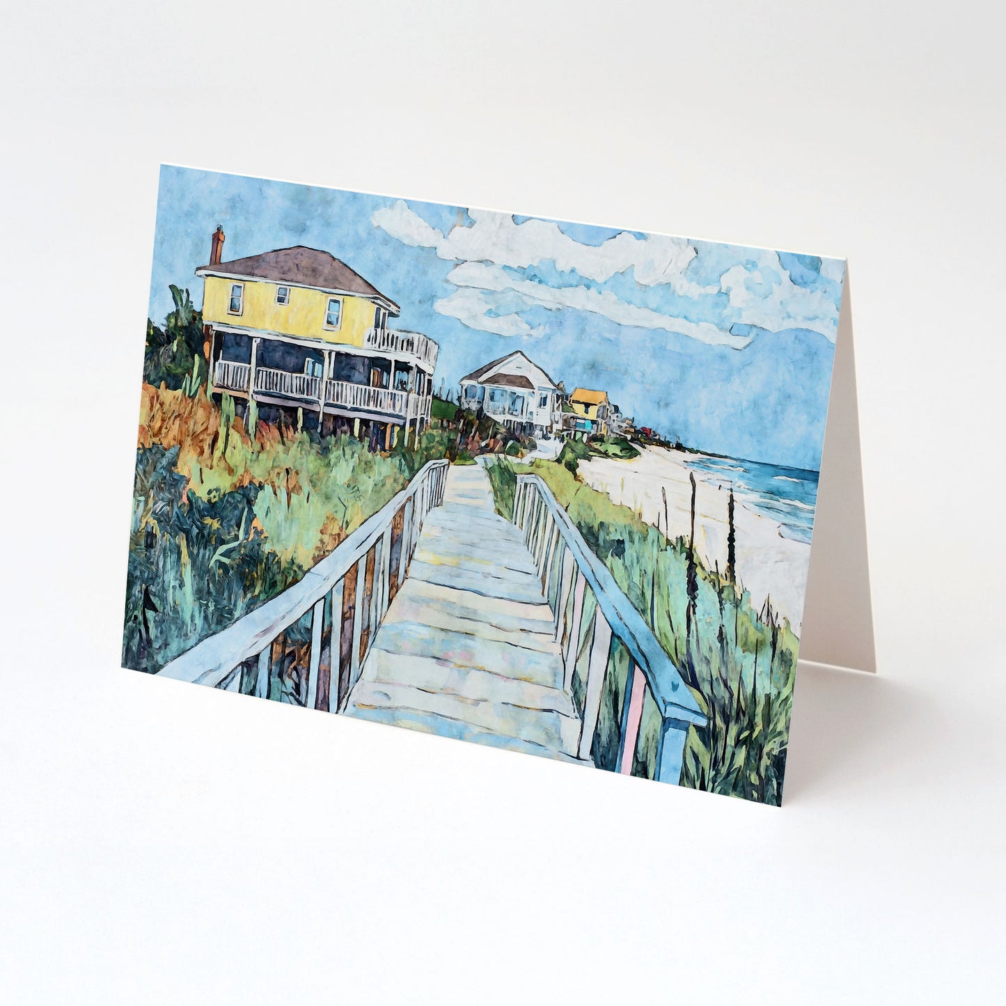A folded greeting card featuring a watercolor-style illustration of a coastal boardwalk. The scene shows a weathered wooden walkway leading past a bright yellow beach house on stilts, with additional seaside cottages and the ocean visible in the background under a blue sky with scattered clouds.