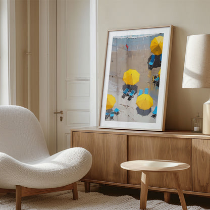 Framed painting of yellow beach umbrellas and blue lounge chairs leaning against a wooden sideboard next to a white textured chair in a well-lit room.