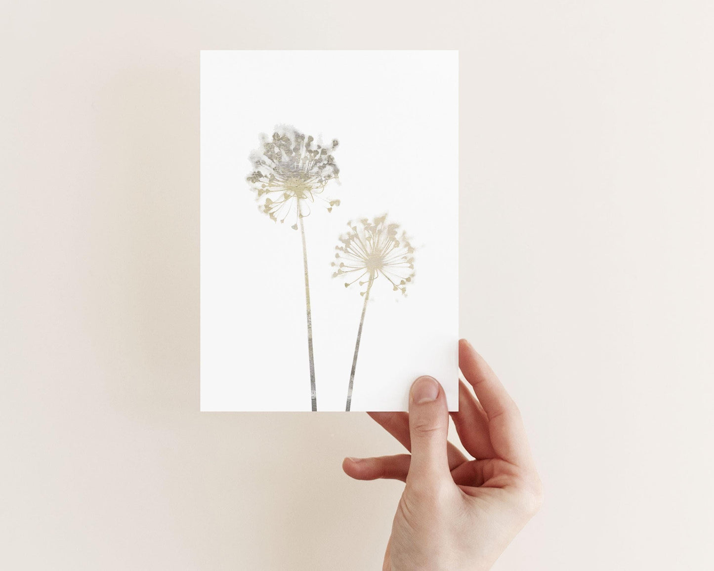 Hand holding a minimalist greeting card with two soft beige watercolor allium flowers on a white background, shown against a pale neutral wall.