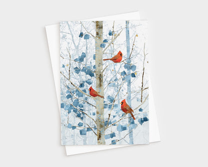 Greeting card featuring three bright red cardinals perched on slender branches of a winter tree. The background shows soft blue and white abstract foliage and a pale birch-like trunk, creating a serene snowy forest scene. Card is displayed on top of a white envelope against a light backdrop.